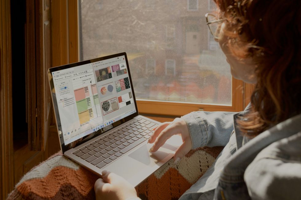a woman sits on a window sill and uses her surface laptop with windows 11